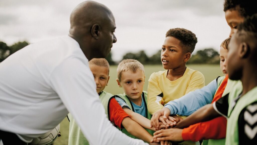 An adult and a group of kids put their hands into the middle of their group in a show of support.