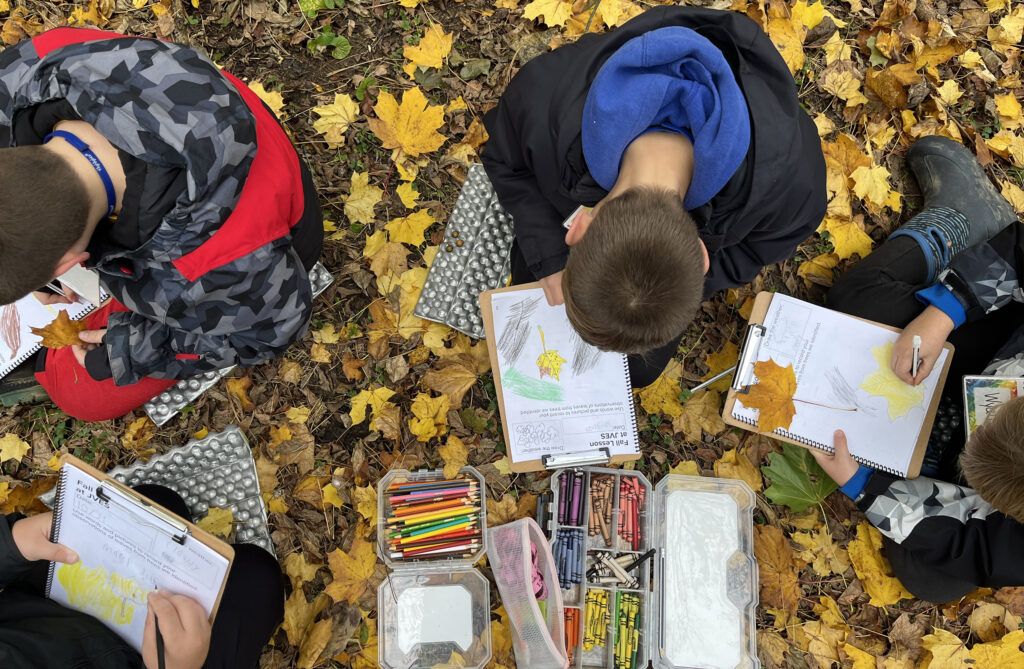 Children sit on the ground among fallen autumn leaves, using clipboards and art supplies to sketch and color leaves as part of an outdoor learning activity.