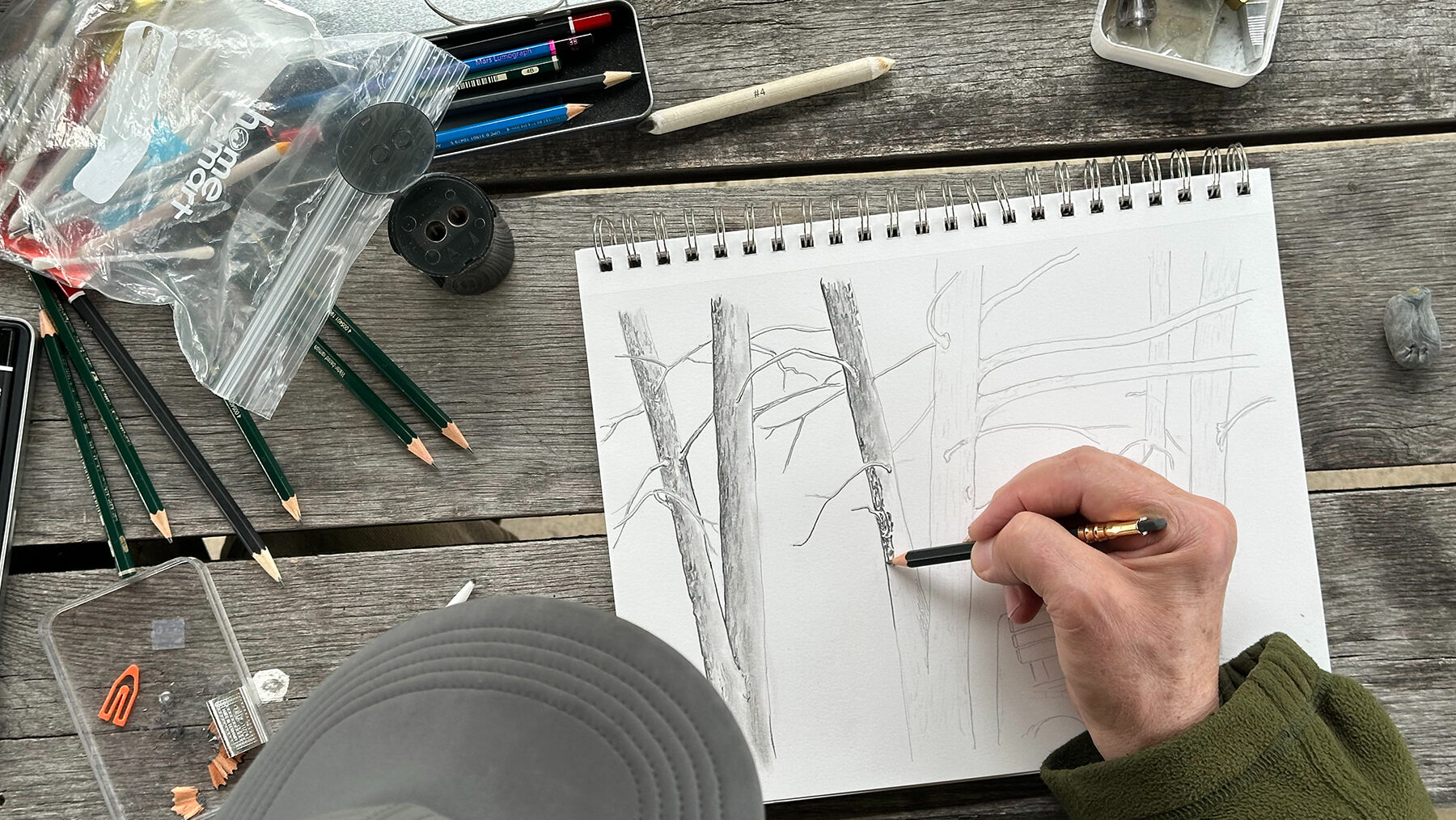 A cozy, outdoor sketching setup on a rustic wooden table, featuring an artist drawing tree trunks in a sketchbook using a pencil. The scene includes scattered art supplies like sharpened pencils, erasers, a pencil sharpener, tape, and a plastic bag of colored markers and tools. The artist, wearing a cap and a warm jacket, is focused on capturing the details of the forest scene, creating a peaceful, nature-inspired artistic moment.