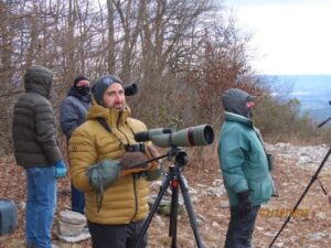 A group of bundled-up birdwatchers, including a man using a spotting scope, observe the sky from a rocky, wooded vantage point on a cold winter day.