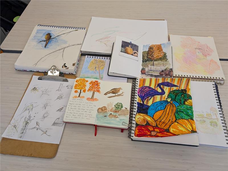 A tabletop display showcasing a collection of sketchbooks and drawings with various nature-themed illustrations. The artwork includes sketches of birds, vibrant fall trees, pumpkins, and other autumn scenery, blending pencil sketches, watercolors, and colorful markers. The mix of nature studies and creative drawings creates a cozy and artistic environment, perfect for exploring seasonal inspiration.